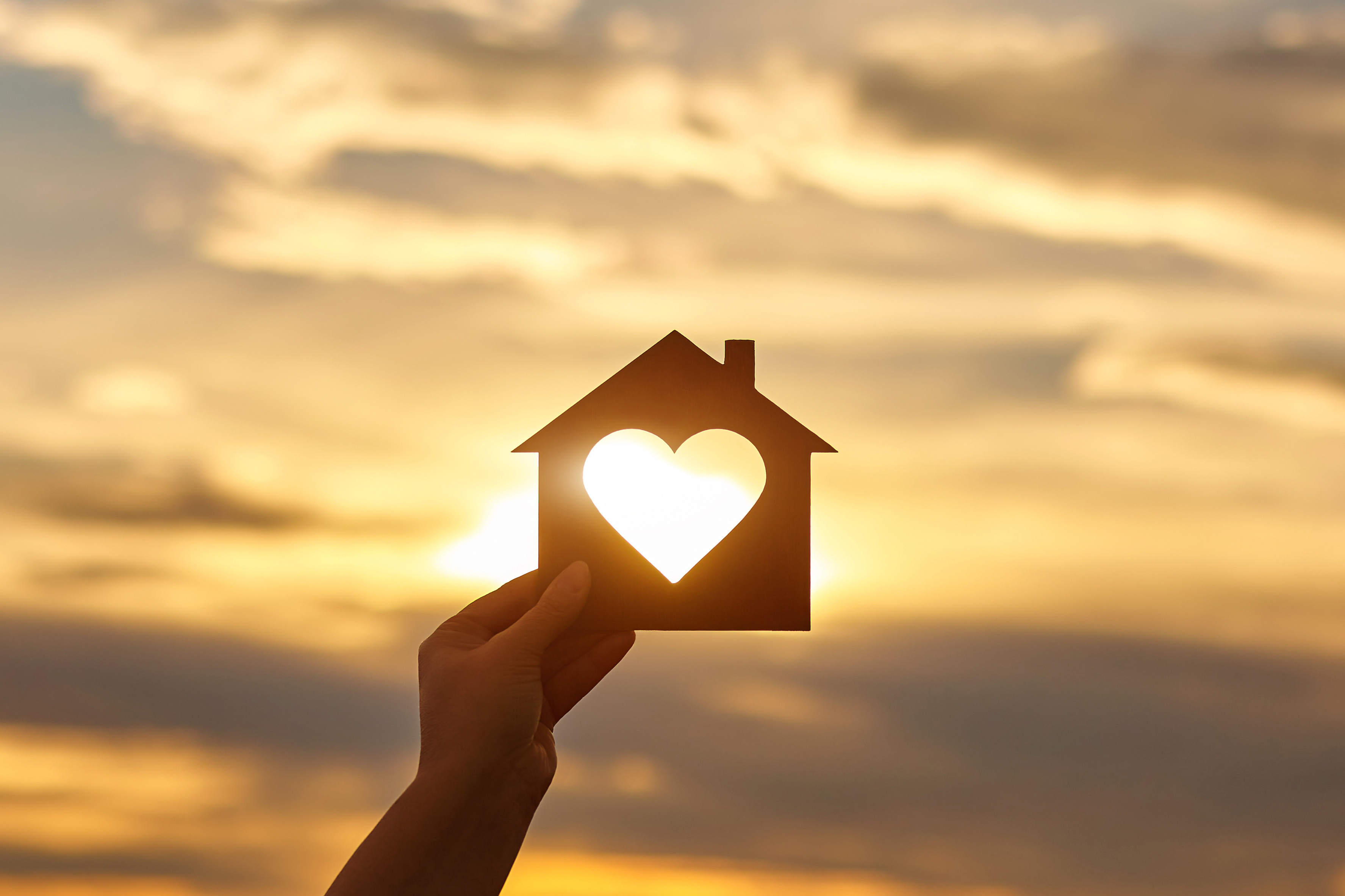 Woman hand holds wooden house in the form of heart against the sunset